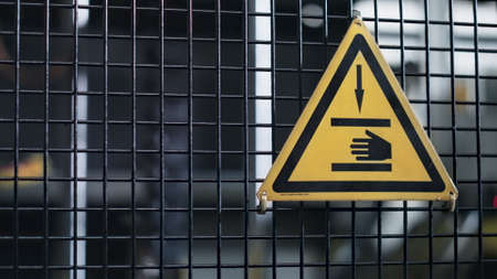 Factory machine danger sign with hand icon on grid conveyor mechanism background. Yellow metal triangle caution symbol in mechanic manufacture attention information. Safety production beware conceptの写真素材