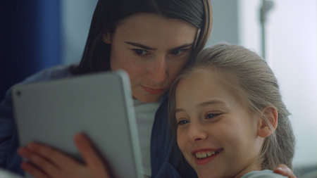 Family enjoy playing tablet computer closeup. Joyful mom kid have fun together. Smiling blond girl holding pad surfing internet with beautiful young mother. Caring parent hug communicate with child.の写真素材