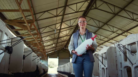 Smiling farmer holding bottle for feeding newborn. Happy agribusiness worker. Joyful woman professional livestock manager posing at feedlots ready to help calves cowshed. Animal husbandry concept.の写真素材