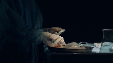 Nurse hands preparing tools for wound suturing in dark operating room closeup. Unrecognized assistant helping surgeon open surgery equipment alone. Healthcare professional work with medical instrumentの写真素材