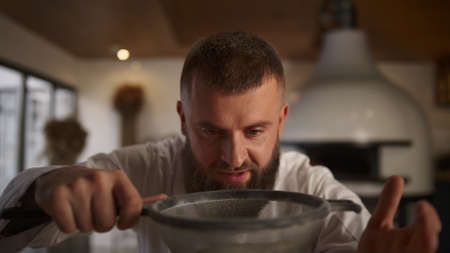 Baker man sifting flour using sieve in culinary restaurant. Close up chef cooking in professional kitchen cafe. Handsome pastry cook making bread pizza dough in dining place. Delicious meal concept.の写真素材