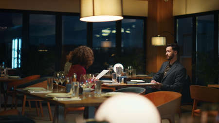 Romantic lovers sitting in night hotel restaurant together. Multiethnic elegant couple spending time on dinner date. Two people looking choosing menu in evening city cafe. Anniversary romance concept.の写真素材