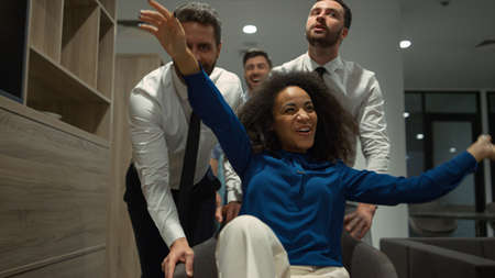 Multi ethnic business team having fun in office. Multiracial company riding chair race in corporate...