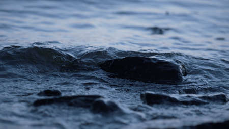 Fast stream flow dark stones evening close up. Cold lake water wash rocks shore on cloudy day....