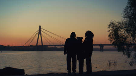 Parents kid silhouette watching sunset standing together at river bridge view. Unknown man father talking with loving family daughter curly wife at clear evening sky. Happy carefree childhood conceptの写真素材