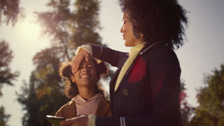 Happy mother kid making selfie together in warm golden sunlight spring park. Smiling african american woman mother hugging carefree daughter hold smartphone outdoors. Loving caring motherhood concept.の写真素材