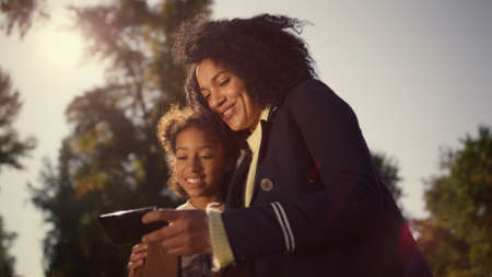 Happy mother daughter selfie posing in golden sunlight. Joyful time together. Attractive african american woman holding smartphone make picture with smiling kid. Cute family enjoying moment sunny dayの写真素材