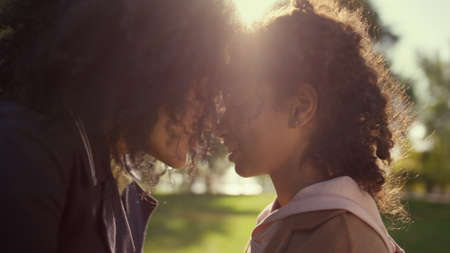 Loving mother touching noses with dreamy curly daughter golden sunlight closeup. Happy african...