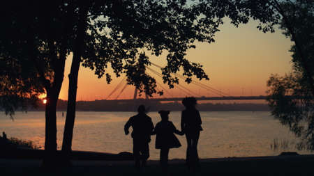 Happy family silhouette run to river hold hands. Beautiful bridge waterway view. Unknown loving...