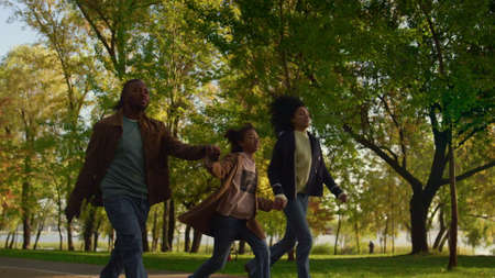 Happy family walking autumn park holding hands evening. Caring people together. Relaxed loving african american parents enjoying outdoor leisure time with smiling daughter. Carefree childhood conceptの写真素材