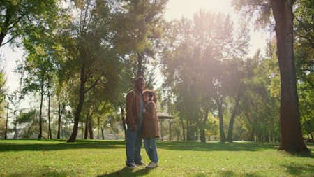 Happy child father standing together on green park field in golden sunlight. Joyful african american man smiling dad hugging cute curly daughter outdoors. Loving family resting enjoy spring warm day.の写真素材