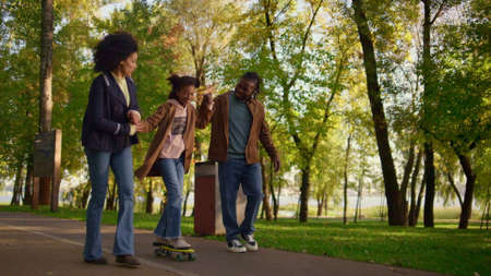 Parents teaching girl riding longboard in park. Supportive family encourage kid. Joyful african american couple holding daughter hands help balancing. Loving father caring mother enjoying parenthood.の写真素材