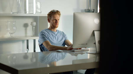 Office worker typing computer keyboard writing email at cozy remote workplace. Focused ginger man...