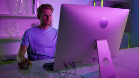 Focused gamer playing video game. Creative architect make project in neon lights. Serious man enjoying online shooter cyber competition on weekend. Redhead guy freelancer using working pc computer.の写真素材