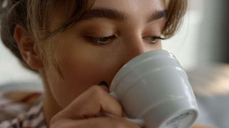 Hand holding coffee cup closeup. Relaxed girl enjoy morning beverage on couch. Attractive blonde wife tasting hot tea resting on breakfast time. Beautiful woman taking sip begin idyllic weekendの写真素材