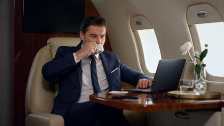 Stressed professional working laptop in airplane. Focused man taking coffee sip in business class...