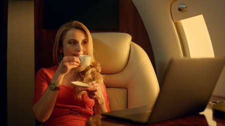 Wealthy woman traveling business airplane closeup. Attractive lady in sunlight drinking coffee....