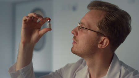 Young physician holding vaccine vial sitting desk hospital office close up. Man doctor examining...