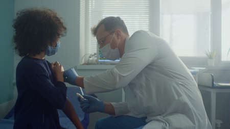 Professional medic injecting covid vaccine to african american teen child close up. Man physician wearing protective face mask vaccinate preteen girl in hospital office. Preventive treatment concept.の写真素材