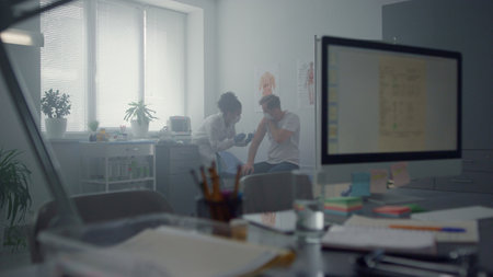Middle age man getting covid vaccination in clinic office. African american woman doctor making injecting antivirus preparate in modern hospital cabinet. Computer screen with vaccine patient data.の写真素材