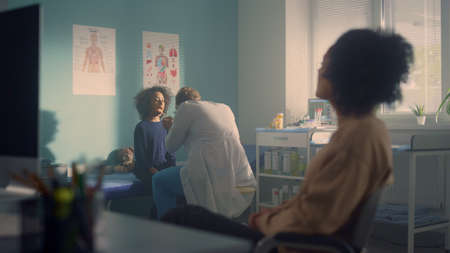 Pediatrician examining girl throat checking on coronavirus symptoms. African mom sitting waiting on...
