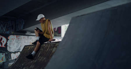 Stylish man browsing internet online on mobile phone at skatepark. Young skater scrolling surfing...