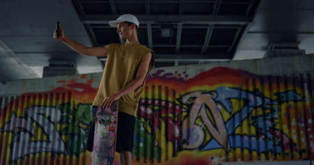 Casual skater taking selfie photo on smartphone at skate park with graffiti wall. Stylish teenager...