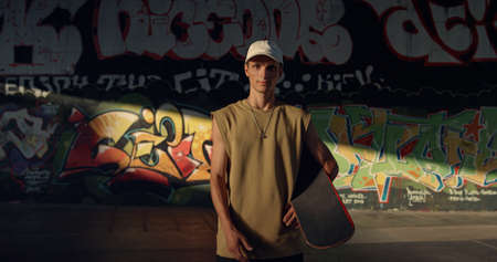 Attractive man skateboarding at urban skate park. Casual active teenager hipster looking on camera at training place with graffiti wall background. Stylish extreme sport urban subculture concept.の写真素材