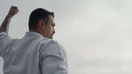 Sporty man doing karate exercises wearing kimono against cloudy gray sky close up. Bearded sportsman practicing martial arts outdoors. Focused athlete training fighting technique on gloomy beach.の写真素材