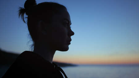 Dark silhouette face woman standing in front blue evening sky close up. Calm girl relaxing on beach...