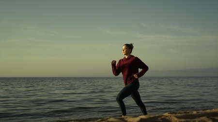 Attractive girl runner training on sandy beach evening time. Sporty woman running near calm ocean...