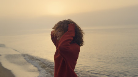 Excited girl jumping on beach. Smiling woman turning around on sea coast. Joyful lady raising hands. Female hipster feeling happy. Stylish woman touching curly hair with hands. Sunrise landscapeの写真素材
