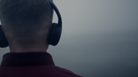 Closeup young guy listening music in headphones on beach. Back view unrecognizable man enjoying music song in earphones. Male hipster moving head in music rhythm. Focus on waving sea in morning mistの写真素材