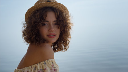 Adorable young woman relax on sunny beach wearing yellow flowery dress close up. Seductive curly girl posing in front calm blue ocean. Portrait of gorgeous beautiful lady in straw hat summer day.の写真素材