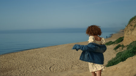 Laughing pretty girl running down seashore with arms outstretched sunny morning. Happy curly woman walking beautiful empty beach. Carefree attractive lady have fun on nature wearing jeans jacket.の写真素材