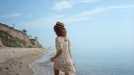 Cute curly girl dancing on ocean waves holding dress in hands. Smiling attractive woman have fun on sunny seashore wearing straw hat summer day. Happy carefree lady raising arms walking on water.の写真素材