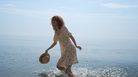 Happy curly woman spinning bouncing on wet sand beach. Energetic cheerful girl wearing cute dress walking on ocean waves holding straw hat. Attractive laughing lady have fun on beautiful summer shore.の写真素材