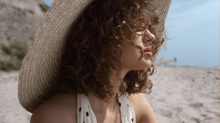 Closeup of sexy slim girl legs on sunny seashore. Beautiful seductive woman wearing wide brimmed hat smearing feet with sunscreen. Attractive curly model in swimsuit sitting sand beach summer weekend.の写真素材
