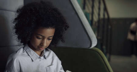Cute african american preteen girl sitting alone on bench close up. Pretty schoolgirl take off backpack after classes in school corridor. Curly creative pupil get out sketchpad to drawing picture.の写真素材
