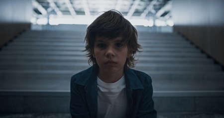 Lonely teen schoolchild sitting alone on school staircase closeup. Unhappy schoolboy posing on stairs looking camera. Elementary age boy hide in empty dark hall. Social bullying victim concept.の写真素材