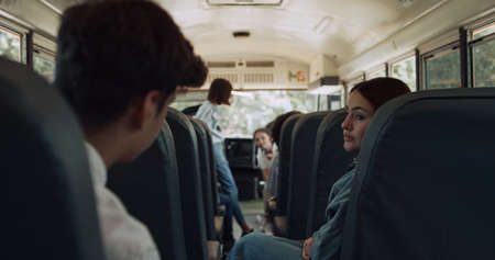 High school students sitting communicating inside schoolbus. Smiling pupils waving to boarding schoolgirl friend. Diverse multi ethnic teen schoolchildren talking in vehicle education transport.の写真素材