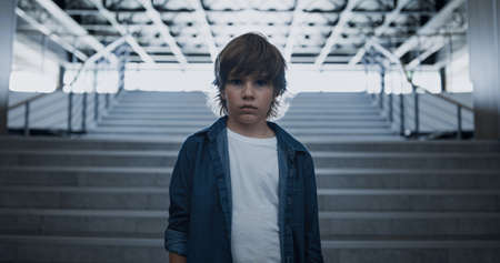 Sad boy face looking camera unhappily close up. Depressed schoolboy standing alone at empty staircase. Upset little outcast feeling despair suffer after class bullying. Loneliness in school concept.の写真素材