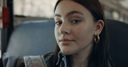 Portrait of beautiful girl looking camera sitting in empty school bus alone. Pensive brunette student posing at vehicle window with backpack. Pretty schoolgirl passenger on daily trip close up.の写真素材