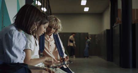 Three focused students sitting using tablet in school hall. Serious interested friends surfing internet touching pad screen on break together. Preteen children holding digital gadget in corridor.の写真素材