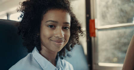 Smiling African American schoolgirl looking camera in school bus close up. Happy joyful girl with curly hair posing at window. Cute brunette pupil enjoying road trip with friend. Childhood concept.の写真素材