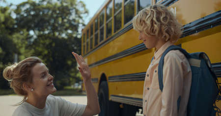 Cheerful mom giving son five at school transport close up. Smiling schoolboy get in yellow...