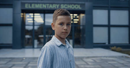 Upset elementary age boy standing at empty schoolyard close up. Portrait of unhappy pupil with sad eyes posing alone at modern campus entrance. Teen child looking camera near school building.の写真素材