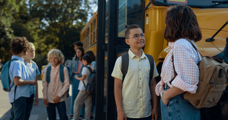 Two joyful pupils stand chatting at schoolbus waiting bus boarding sunny morning. Cute friends smiling talking near academic yellow shuttle. Elementary age boy in glasses communicating with happy girlの写真素材