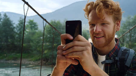 Cheerful hiker make video on cell phone. Close up ginger backpacker enjoy travel blog on river bridge. Happy tourist taking photo using mobile device in mountains. Travel modern holiday concept.の写真素材