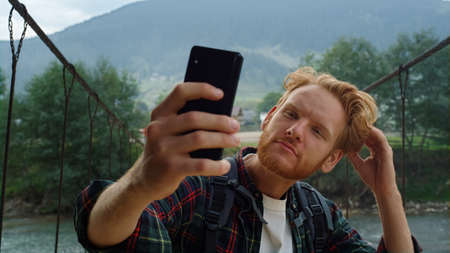 Confident hipster photographing selfie photo using mobile phone on hike travel. Handsome ginger tourist posing camera on smartphone close up. Stylish man fix hair on nature. Modern holiday concept.の写真素材